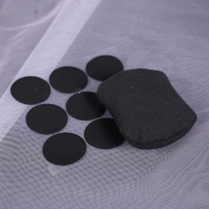 May include: A collection of black circular fabric patches and a small, black rectangular fabric object. The items are arranged on a sheer, white fabric background. The patches are evenly spaced, and the rectangular object is slightly rounded.
