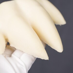 May include: A cream-colored, three-pronged claw-like object with a smooth, matte surface. The structure features three distinct, pointed extensions that curve slightly, presenting a solid, uniform appearance throughout the entire piece.