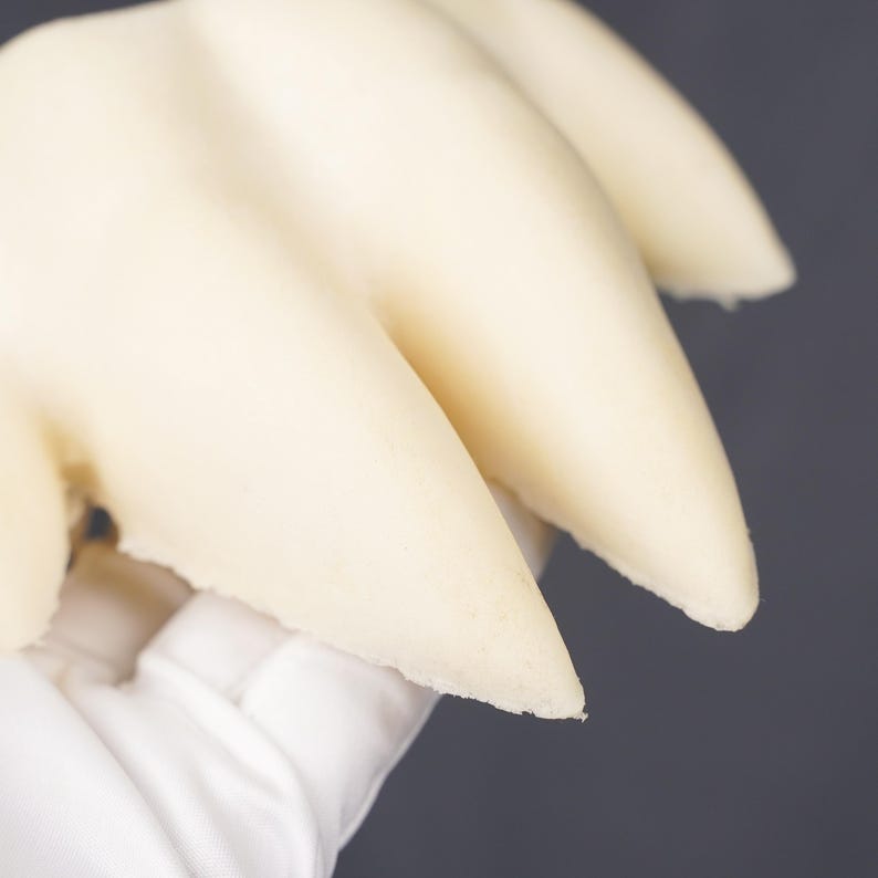 May include: A cream-colored, three-pronged claw-like object with a smooth, matte surface. The structure features three distinct, pointed extensions that curve slightly, presenting a solid, uniform appearance throughout the entire piece.