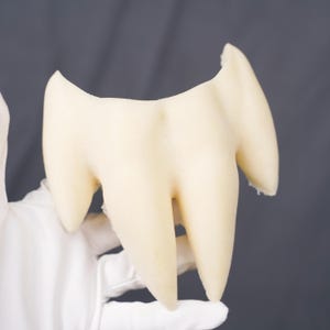 May include: A cream-colored, three-pronged tooth model featuring a smooth, matte surface. The object has a wide, curved top section that tapers down into three distinct, pointed roots, mimicking the anatomical structure of a human molar.