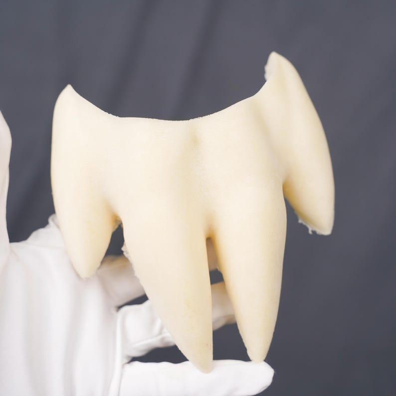May include: A cream-colored, three-pronged tooth model featuring a smooth, matte surface. The object has a wide, curved top section that tapers down into three distinct, pointed roots, mimicking the anatomical structure of a human molar.