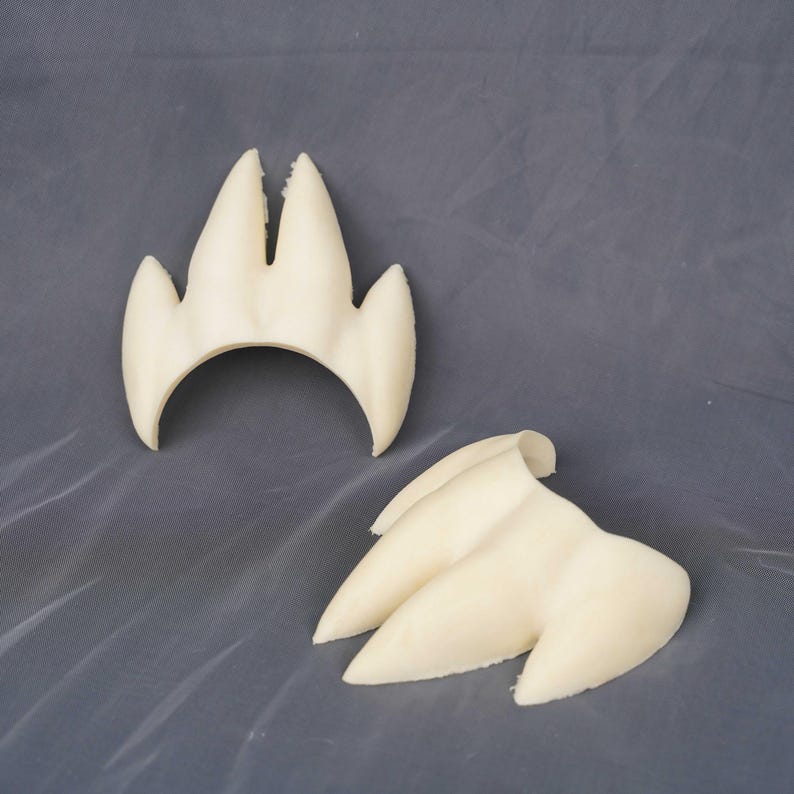 May include: Two off-white, three-pronged claw-like objects with a smooth, matte finish. Each piece features a curved base and three distinct, pointed extensions that resemble animal talons or claws, presented as separate, individual components.