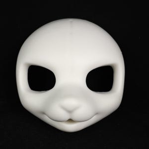 May include: A blank, white doll head with large, oval eye sockets and a small, rounded nose. The head has a smooth surface and a slight smile, suggesting a friendly or neutral expression. The doll head is likely for customization.