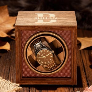 Pode incluir: Um enrolador de relógios de madeira com um relógio dentro. O enrolador é quadrado com um acabamento castanho e uma janela de visualização de vidro. O relógio tem um mostrador castanho e uma bracelete prateada. A parte superior do enrolador tem o nome "BRIAN" gravado.