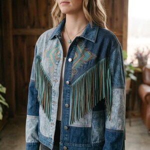 May include: A denim jacket featuring a patchwork design of light and dark blue acid-wash panels. The front displays a geometric embroidered pattern in teal and gold, accented by long, hanging fringe in matching teal and brown tones.