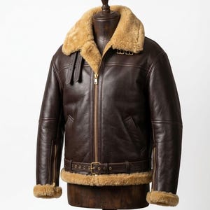 Peut inclure: Veste de bombardier en cuir marron avec col et doublure en peau de mouton beige. La veste est dotée d'une fermeture éclair sur le devant, d'une ceinture à la taille et de bordures en peau de mouton aux poignets. La veste est présentée sur un mannequin en bois.