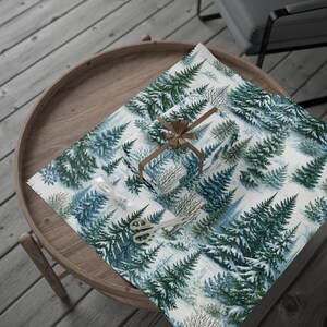 May include: A rectangular gift box wrapped in white paper featuring a repeating pattern of dark green pine trees covered in snow. The box is tied with a brown ribbon in a bow. A pair of metal scissors and a clear tape dispenser are placed nearby.