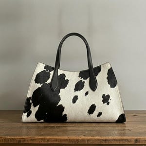 Structured wide-format cowhide tote bag in bold black and white cow print hair-on hide, short dual tapered black leather top handles, open-top trapeze silhouette, self-supporting, photographed front-facing on warm reclaimed wood table against grey wall.