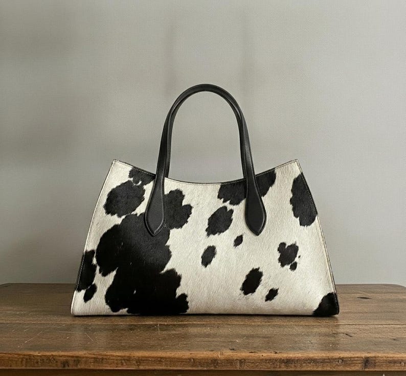 Structured wide-format cowhide tote bag in bold black and white cow print hair-on hide, short dual tapered black leather top handles, open-top trapeze silhouette, self-supporting, photographed front-facing on warm reclaimed wood table against grey wall.