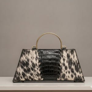Può includere: Una borsa a mano trapezoidale con un motivo a pelle di mucca bianco e nero sui lati e un pannello centrale verticale con una texture nera lucida in rilievo. La borsa include un singolo manico rotondo in metallo dorato sul telaio superiore.