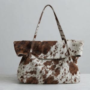 May include: A tote bag featuring a brown and white cowhide pattern with a textured fur finish. The bag has a wide, folded top opening and two long, thin shoulder straps that extend from the front and back panels of the main body.