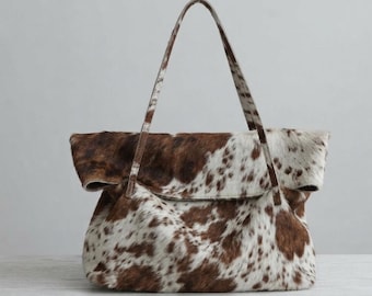 Può includere: Una borsa tote con motivo a pelle di mucca marrone e bianca e finitura in pelliccia testurizzata. La borsa presenta un'ampia apertura superiore ripiegata e due lunghi spallacci sottili che si estendono dai pannelli anteriore e posteriore.