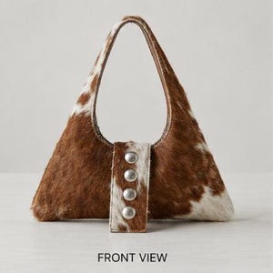 Brown and white cowhide hobo bag with curved shoulder strap and pearl snap button trim, hair-on-hide leather purse for western boho style, shown on a light wood tabletop.