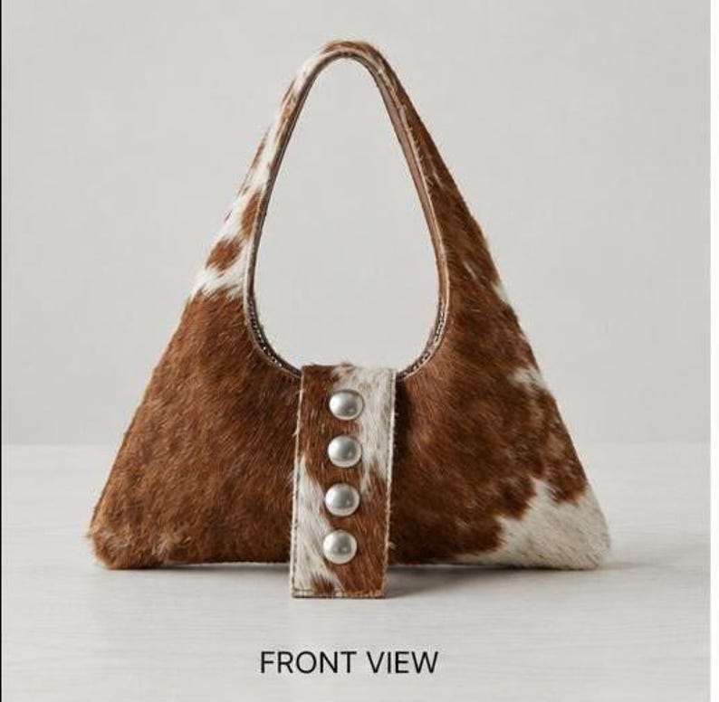 Brown and white cowhide hobo bag with curved shoulder strap and pearl snap button trim, hair-on-hide leather purse for western boho style, shown on a light wood tabletop.
