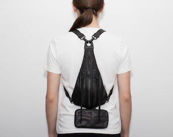 May include: Black leather backpack with a distinctive triangular shape, a central zip, and several small pockets. It has adjustable straps and a small, rectangular pouch. The backpack is worn over a white t-shirt. The bag is worn over a white t-shirt.