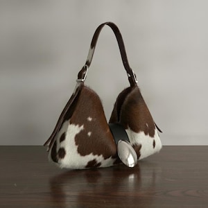 Brown and white speckled cowhide triangular hobo bag with a single dark leather strap, polished silver disc clasp, and fringe tassels, displayed on a dark wood table against a soft grey backdrop.