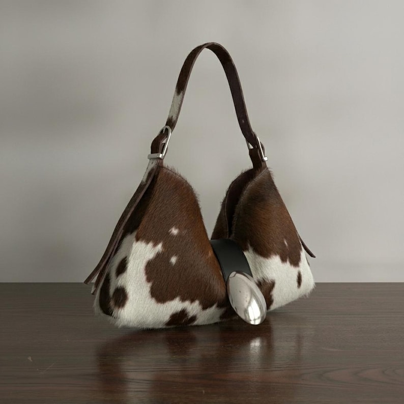 Brown and white speckled cowhide triangular hobo bag with a single dark leather strap, polished silver disc clasp, and fringe tassels, displayed on a dark wood table against a soft grey backdrop.