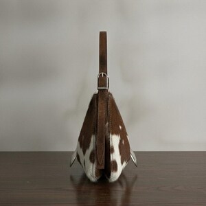 Brown and white speckled cowhide triangular hobo bag with a single dark leather strap, polished silver disc clasp, and fringe tassels, displayed on a dark wood table against a soft grey backdrop.