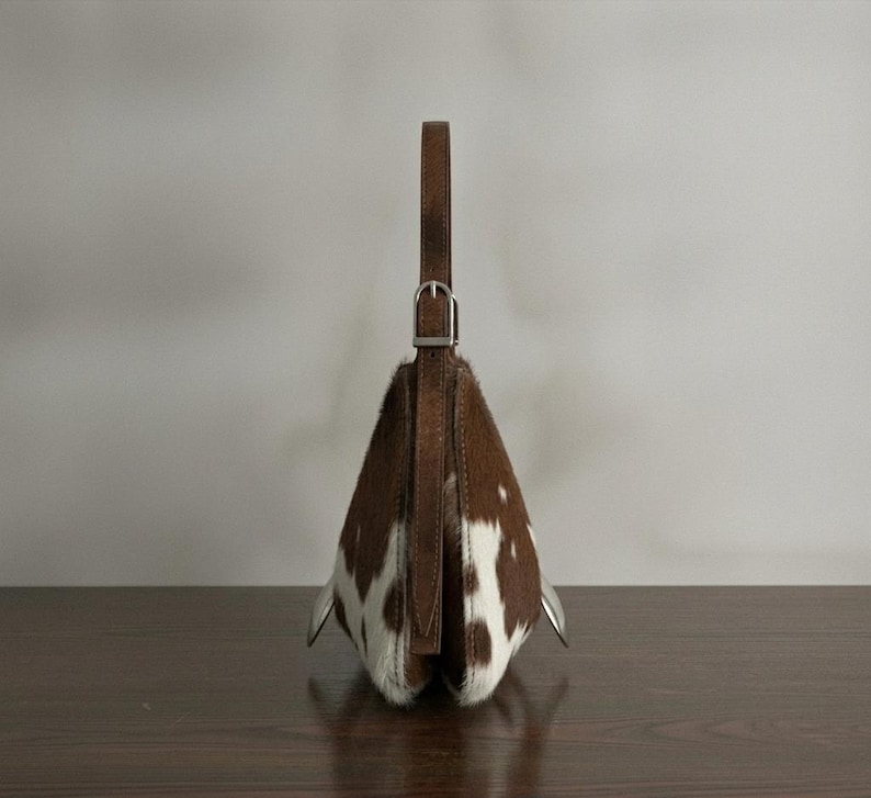 Brown and white speckled cowhide triangular hobo bag with a single dark leather strap, polished silver disc clasp, and fringe tassels, displayed on a dark wood table against a soft grey backdrop.