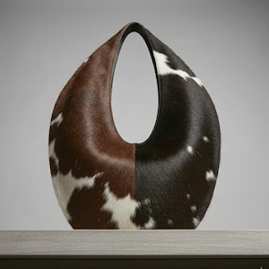 Oval teardrop loop shoulder bag made from genuine tricolor hair-on cowhide, showing brown and dark chocolate panels with natural white markings, photographed standing upright on a grey wooden surface against a light grey wall.