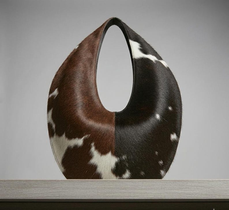 Oval teardrop loop shoulder bag made from genuine tricolor hair-on cowhide, showing brown and dark chocolate panels with natural white markings, photographed standing upright on a grey wooden surface against a light grey wall.