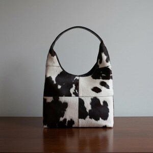 May include: A hobo-style handbag constructed from panels of black and white cowhide featuring a natural hair-on texture. The bag has a single arched top handle and a structured, rectangular base with a distinct patchwork design of animal print.