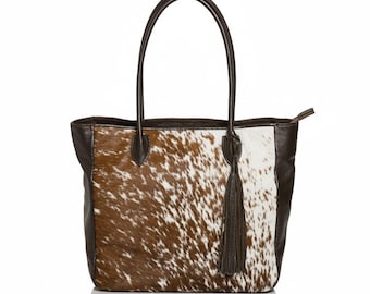 May include: A brown and white cowhide tote bag with dark brown leather trim and handles. The bag has a long, dark brown tassel and a zip closure. The bag is designed for carrying items.