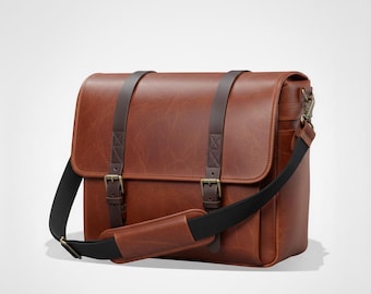May include: A brown leather messenger bag with a flap closure, two buckle straps, and a black shoulder strap. The bag has a rectangular shape and a handle on the side. The bag is made of a smooth, textured leather.