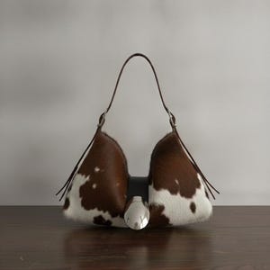 Brown and white speckled cowhide triangular hobo bag with a single dark leather strap, polished silver disc clasp, and fringe tassels, displayed on a dark wood table against a soft grey backdrop.