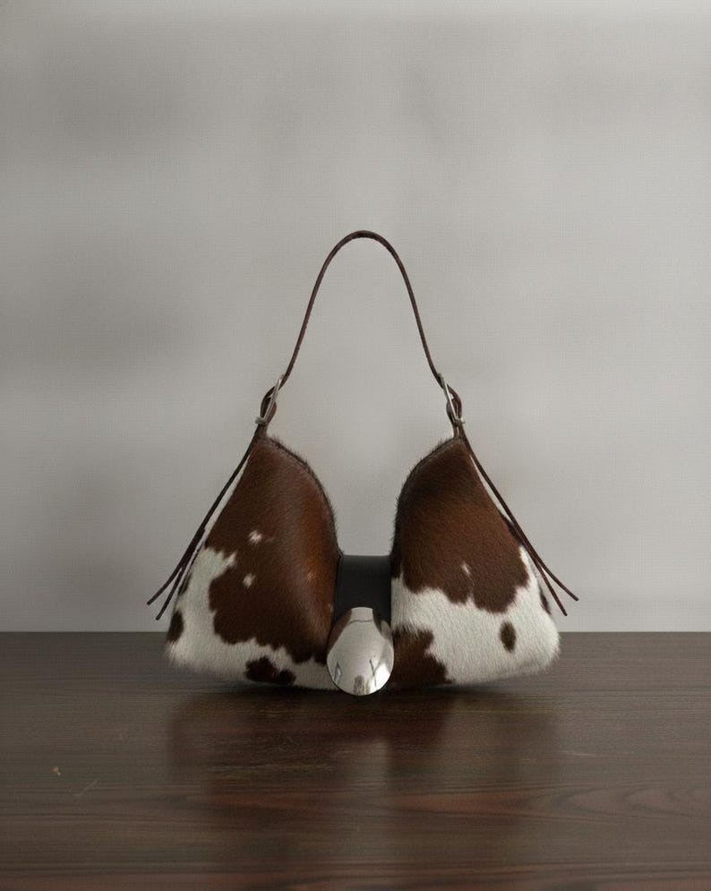 Brown and white speckled cowhide triangular hobo bag with a single dark leather strap, polished silver disc clasp, and fringe tassels, displayed on a dark wood table against a soft grey backdrop.