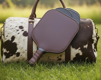 May include: A cowhide leather duffel bag with brown leather handles and accents, alongside two pickleball paddles. The paddles are dark brown with black handles. The bag is on green grass.
