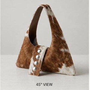 Brown and white cowhide hobo bag with curved shoulder strap and pearl snap button trim, hair-on-hide leather purse for western boho style, shown on a light wood tabletop.