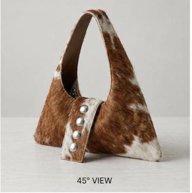Brown and white cowhide hobo bag with curved shoulder strap and pearl snap button trim, hair-on-hide leather purse for western boho style, shown on a light wood tabletop.