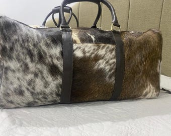 May include: A large, rectangular duffel bag with a cowhide exterior. The bag features a mix of brown, white, and grey fur patterns. It has two dark brown leather handles and a matching shoulder strap. The bag is designed for travel.