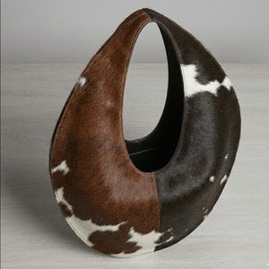 Oval teardrop loop shoulder bag made from genuine tricolor hair-on cowhide, showing brown and dark chocolate panels with natural white markings, photographed standing upright on a grey wooden surface against a light grey wall.
