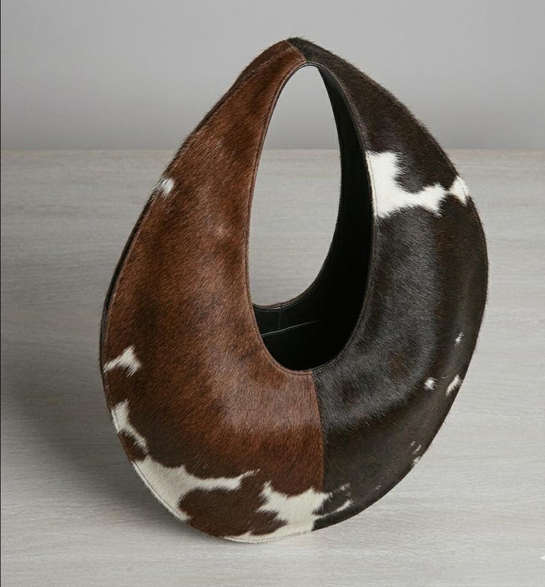 Oval teardrop loop shoulder bag made from genuine tricolor hair-on cowhide, showing brown and dark chocolate panels with natural white markings, photographed standing upright on a grey wooden surface against a light grey wall.