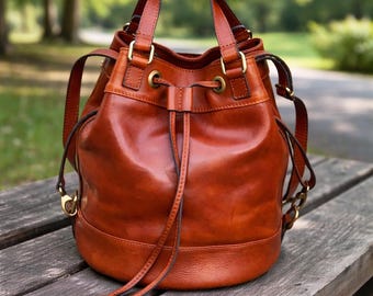 May include: A brown leather bucket bag with a drawstring closure and top handles. The bag features gold-coloured hardware and a rounded base. The bag is on a wooden bench in an outdoor setting.