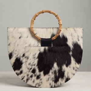 May include: A semi-circular handbag featuring a black and white cowhide pattern with a textured hair-on-hide finish. The bag includes a circular bamboo handle attached by a black leather tab centered at the top edge of the curved body.
