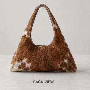 Brown and white cowhide hobo bag with curved shoulder strap and pearl snap button trim, hair-on-hide leather purse for western boho style, shown on a light wood tabletop.