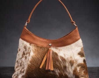 May include: A brown and white cowhide shoulder bag with a leather handle and trim. The bag features a tassel and a silver-tone metal accent. The bag is a crescent shape.