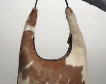 May include: A brown and white cowhide shoulder bag with a curved shape and a dark brown handle. The bag has a zip closure and a soft, textured appearance. The bag is held by a person.