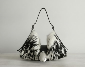Può includere: Una borsa con due pannelli laterali in pelliccia con stampa maculata bianca e nera, collegati da un cinturino centrale in pelle nera con una grande fibbia metallica ovale e liscia color argento e una sottile tracolla nera.