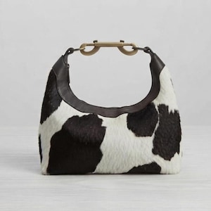 May include: A hobo-style handbag featuring a black and white cow-print pattern on a textured hair-on-hide material. The bag has a dark brown leather trim along the top opening and a metallic brass-toned hardware clasp connecting the handle.