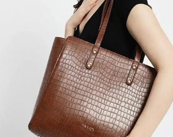 May include: A brown tote bag with a crocodile skin effect. The bag features two handles and gold-coloured rivets. The text "THE CUTIE" is visible on the front.