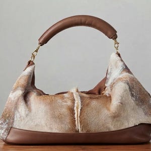 May include: A hobo-style handbag featuring a textured, multi-tonal brown and white animal hide body with a central vertical seam. The bag includes a smooth brown leather base and a matching arched leather handle attached with gold-tone metal clips.