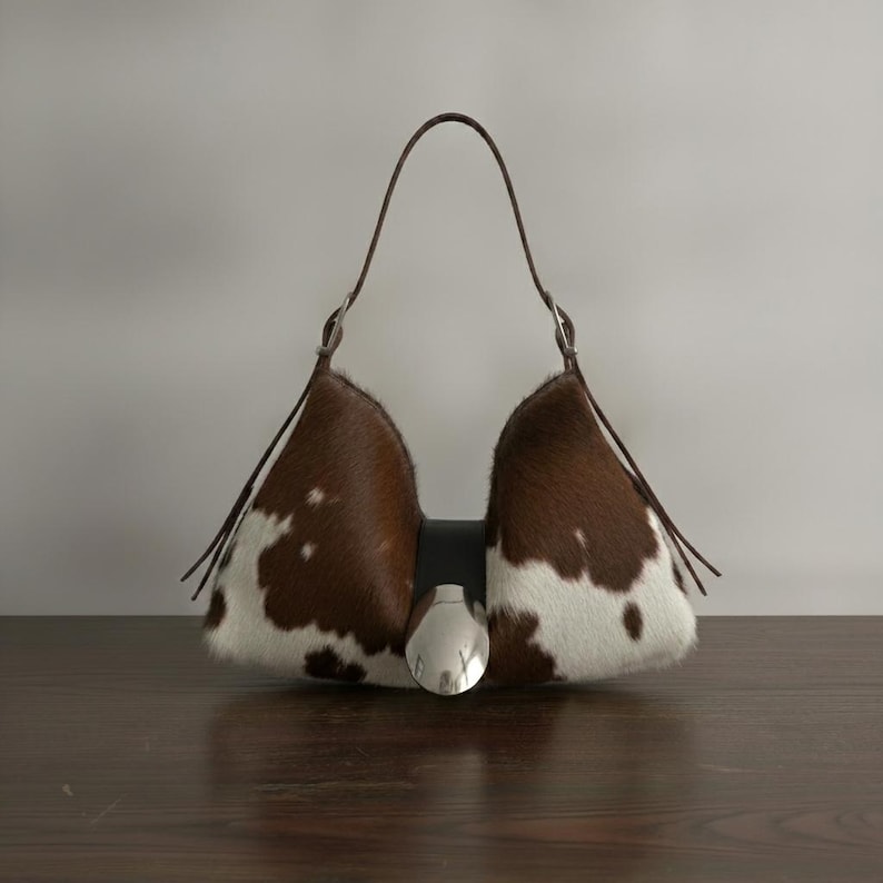Brown and white speckled cowhide triangular hobo bag with a single dark leather strap, polished silver disc clasp, and fringe tassels, displayed on a dark wood table against a soft grey backdrop.