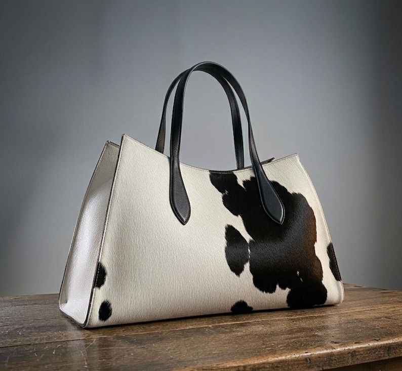 Structured wide-format cowhide tote bag in bold black and white cow print hair-on hide, short dual tapered black leather top handles, open-top trapeze silhouette, self-supporting, photographed front-facing on warm reclaimed wood table against grey wall.