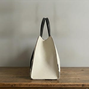 Structured wide-format cowhide tote bag in bold black and white cow print hair-on hide, short dual tapered black leather top handles, open-top trapeze silhouette, self-supporting, photographed front-facing on warm reclaimed wood table against grey wall.