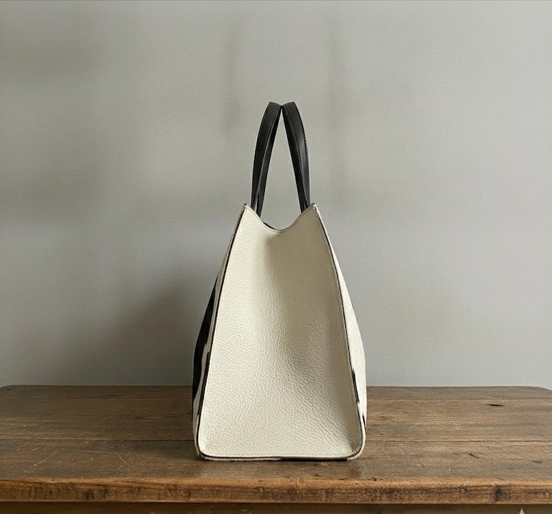 Structured wide-format cowhide tote bag in bold black and white cow print hair-on hide, short dual tapered black leather top handles, open-top trapeze silhouette, self-supporting, photographed front-facing on warm reclaimed wood table against grey wall.