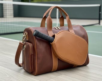 May include: A brown leather duffel bag with a matching brown leather pickleball paddle. The bag has a shoulder strap and a small zipped pocket. The paddle is black and brown.